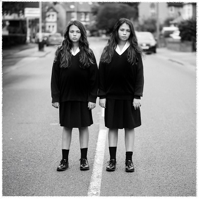 A black and white photo of twin young girls not smiling in school uniform standing in the middle of the road by Cheryl Fisher